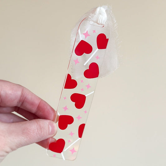 A white hand holds an acrylic bookmark with red hearts attached to white lollipop sticks against a beige wall.